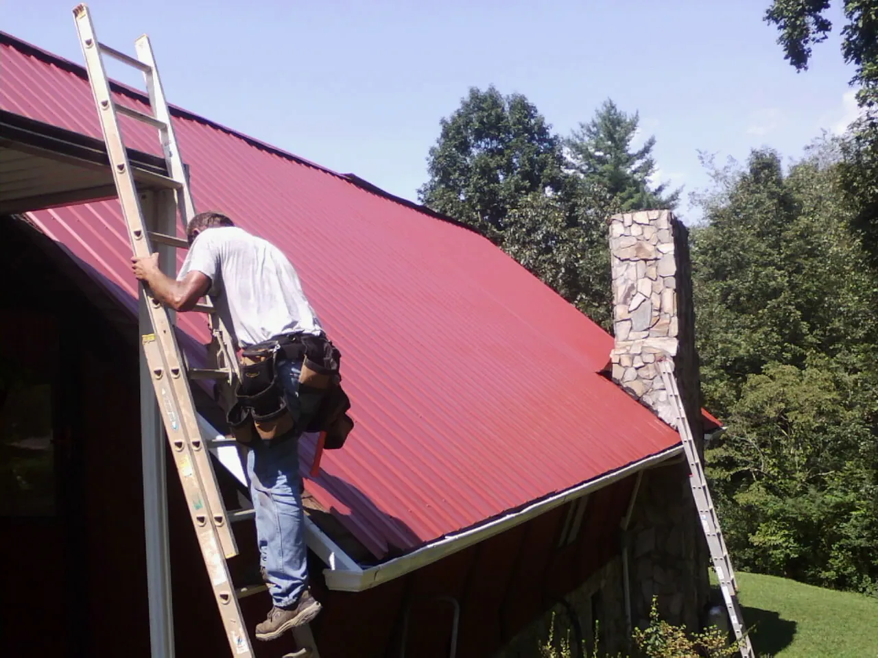 Roofer on a ladder beside a red metal roof and stone chimney — Soffit & Fascia Repair by Roofing Brothers in Du Bois